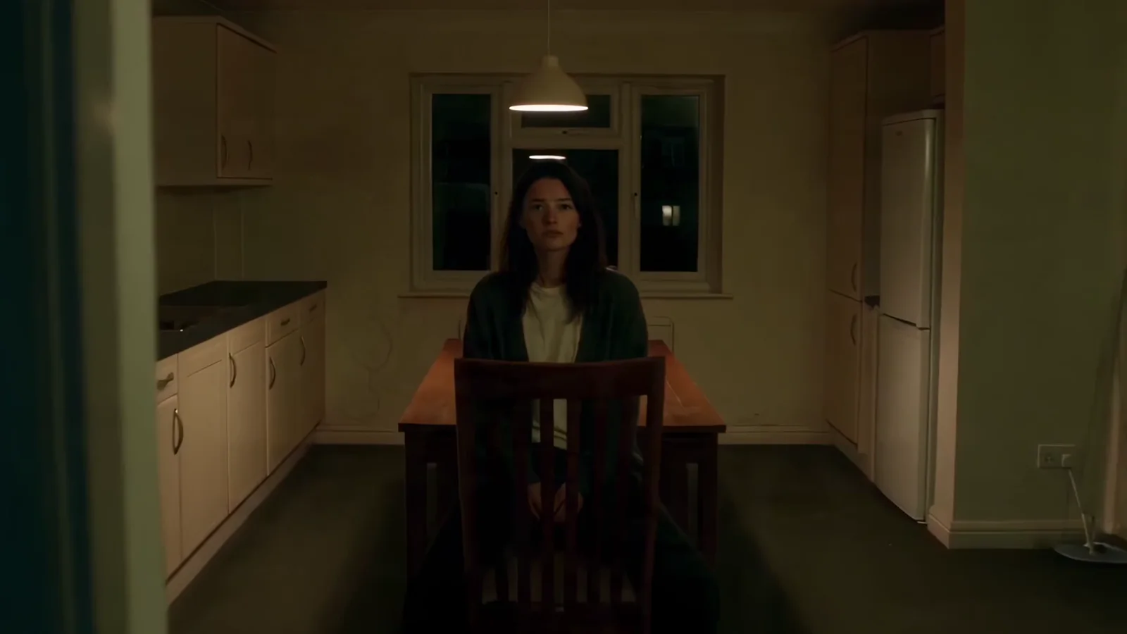 Mara sits alone at a table in a dim kitchen